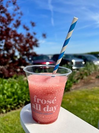 Pink frozen frosé slush in a clear plastic cup labeled "frosé all day" with a blue-and-white striped straw, sitting outdoors on a sunny lawn with parked cars and blue sky in the background.
