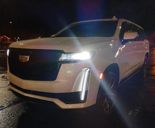 White Cadillac SUV front three-quarter view parked on wet pavement at night with bright LED headlights and vertical daytime running lights reflecting in the lot