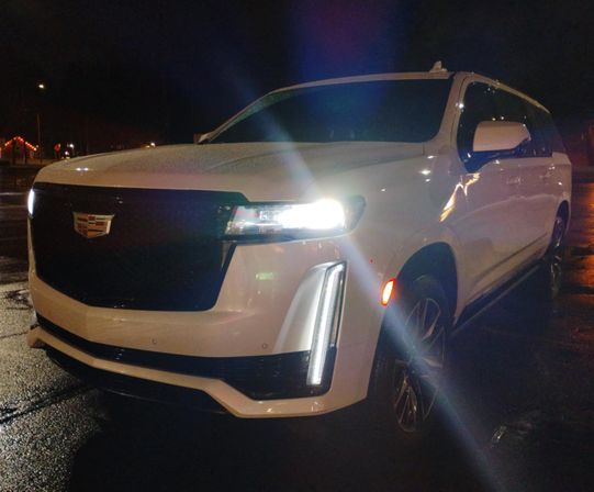 White Cadillac SUV front three-quarter view parked on wet pavement at night with bright LED headlights and vertical daytime running lights reflecting in the lot