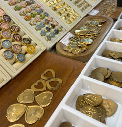 Close-up of a jewelry display at an artisan market: colorful enamel signet rings and gold heart-shaped pendants and initial medallions arranged in trays and wooden dishes.