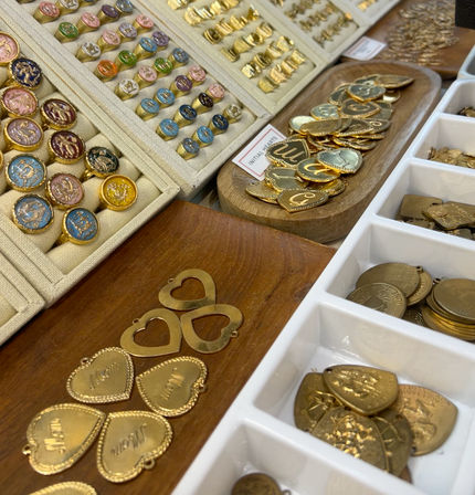 Close-up of a jewelry display at an artisan market: colorful enamel signet rings and gold heart-shaped pendants and initial medallions arranged in trays and wooden dishes.