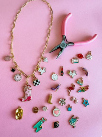 DIY jewelry flatlay on pink background: gold chain necklace with cowboy boot and checkered purse charms, pink-handled pliers and scattered colorful enamel charms (flamingo, heart, star, shoe)
