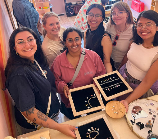 Six smiling women gathered in a bright boutique, holding wooden trays of charm bracelets, chains and tiny earrings over a jewelry display while shopping together.