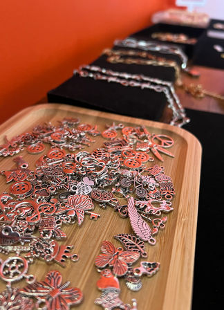 Close-up of silver-tone jewelry charms—butterflies, wings, buttons, bicycles and peace signs—scattered on a bamboo tray with blurred chain necklaces on black displays and an orange backdrop, artisan jewelry display.