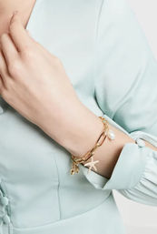 Close-up of a wrist wearing a gold beach-inspired charm bracelet with starfish and pearl charms, styled with a mint-green blouse.