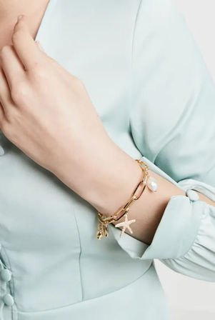 Close-up of a wrist wearing a gold beach-inspired charm bracelet with starfish and pearl charms, styled with a mint-green blouse.