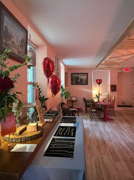 Cozy pink-walled boutique interior with artisan jewelry displayed on a table, shiny red heart-shaped balloons, wooden floors, small seating and framed artwork.