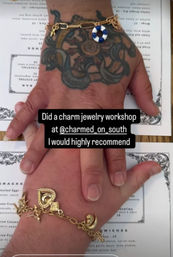 Stacked hands displaying gold charm bracelets—one tattooed wrist with a blue-and-white lifebuoy charm, the other with heart and crescent charms—resting on a printed menu with overlay text praising a charm jewelry workshop.