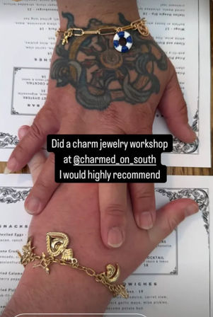 Stacked hands displaying gold charm bracelets—one tattooed wrist with a blue-and-white lifebuoy charm, the other with heart and crescent charms—resting on a printed menu with overlay text praising a charm jewelry workshop.