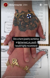 Two hands stacked on a menu after a charm jewelry workshop — a tattooed hand wearing a gold chain with a blue-and-white lifebuoy charm and a second hand wearing chunky gold heart and crescent charms.