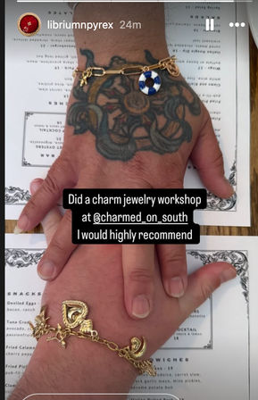 Two hands stacked on a menu after a charm jewelry workshop — a tattooed hand wearing a gold chain with a blue-and-white lifebuoy charm and a second hand wearing chunky gold heart and crescent charms.
