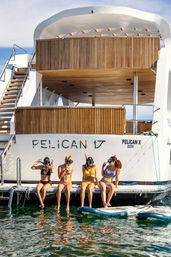 Four women in colorful bikinis wearing snorkeling masks sit on the swim platform of a modern white yacht with teak decks, dangling their feet over sunlit green water beside inflatable paddleboards.