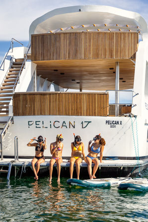 Four women in colorful bikinis wearing snorkeling masks sit on the swim platform of a modern white yacht with teak decks, dangling their feet over sunlit green water beside inflatable paddleboards.