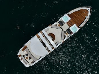 Aerial shot of a luxury yacht on deep blue sea with wooden foredeck, curved sun lounge areas, two jacuzzi tubs, and a swimmer nearby.