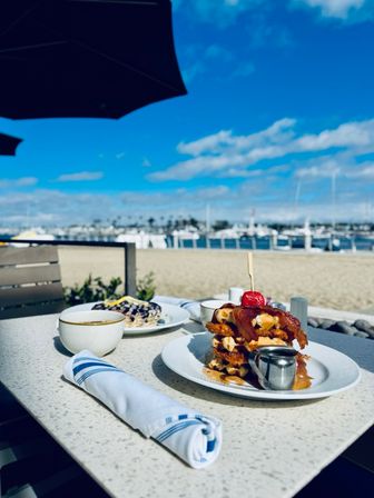 Beachside patio breakfast with stacked waffles topped with bacon and a cherry, syrup pitcher and coffee on a table overlooking a marina and blue sky.