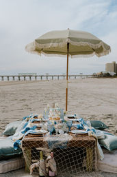 Seaside picnic under a fringed beach umbrella with a low wooden table, blue glassware, cushions and champagne on the sand, pier and ocean in the background.