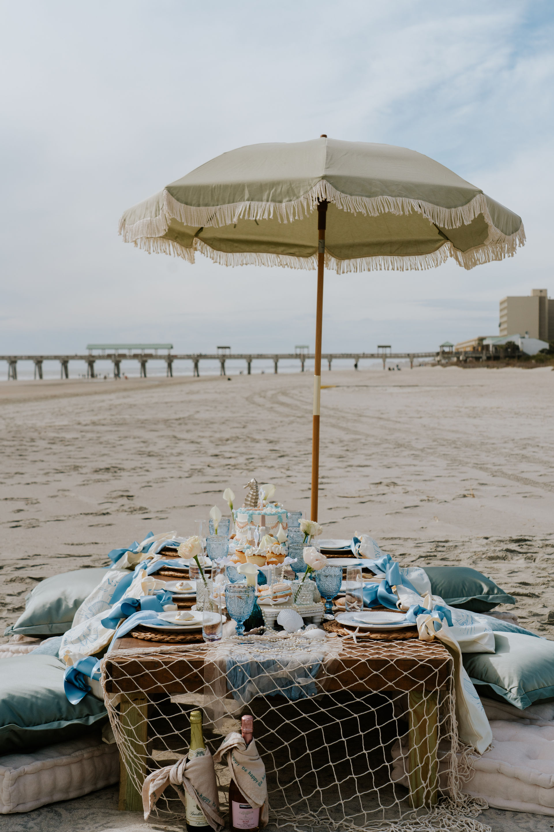 Seaside picnic under a fringed beach umbrella with a low wooden table, blue glassware, cushions and champagne on the sand, pier and ocean in the background.