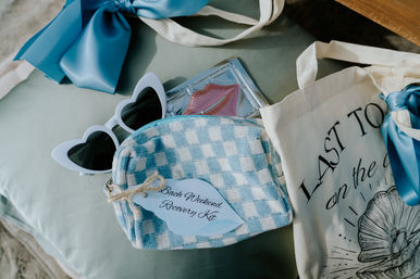 Pastel blue bachelorette flatlay for a beach weekend: gingham recovery kit pouch labeled Bach Weekend Recovery Kit, white heart sunglasses, canvas tote and blue satin bow