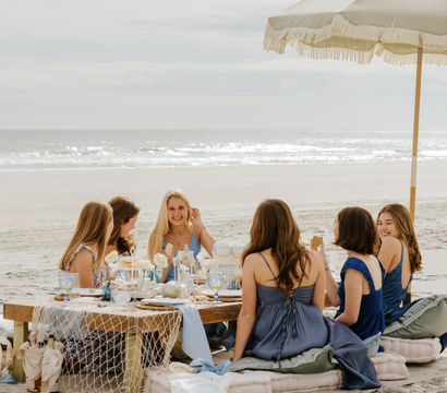 Friends enjoying a relaxed beach picnic under a fringed umbrella on a sandy shore, low table set with desserts and blue accents and the ocean waves on the horizon.