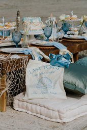 Beachside bridal shower table with pastel frosted cake and cupcakes, blue vintage goblets, seashell accents, woven chargers and a canvas coastal tote on floor cushions.