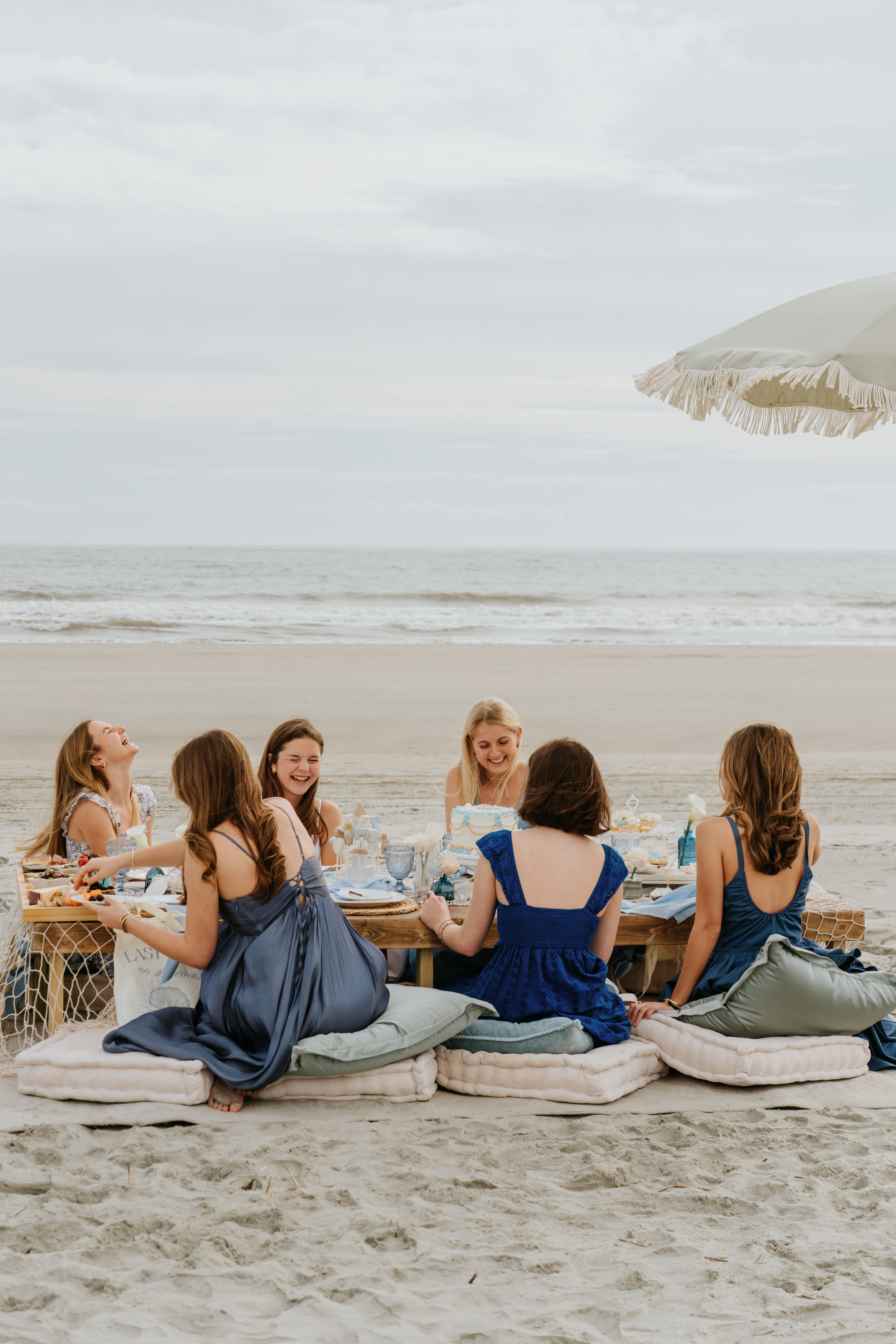 Seaside beach picnic with friends in blue dresses laughing around a low table set with cake, cushions and a fringed umbrella by the ocean