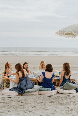 Seaside beach picnic with friends in blue dresses laughing around a low table set with cake, cushions and a fringed umbrella by the ocean
