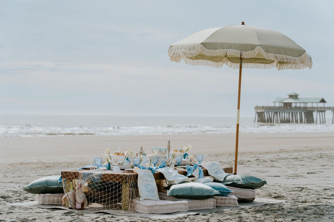 Coastal beach picnic with a fringed umbrella shading a low wooden table set with pastel blue glassware, cushions and decor on the sand, gentle ocean waves and a pier in the background.