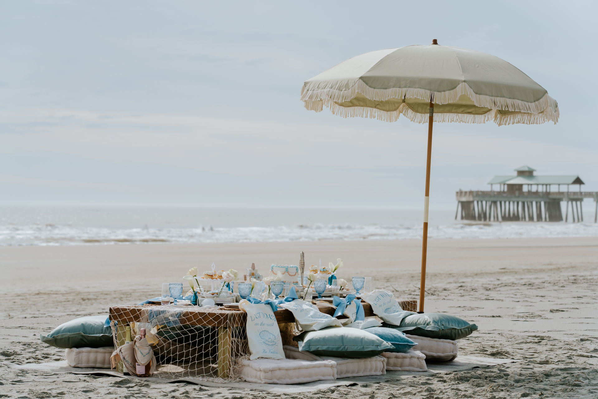 Coastal beach picnic with a fringed umbrella shading a low wooden table set with pastel blue glassware, cushions and decor on the sand, gentle ocean waves and a pier in the background.