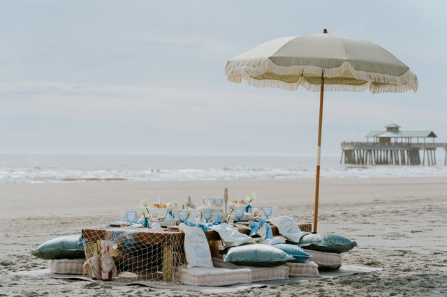 Coastal beach picnic with a fringed umbrella shading a low wooden table set with pastel blue glassware, cushions and decor on the sand, gentle ocean waves and a pier in the background.