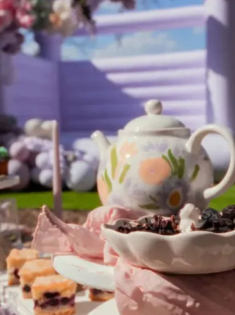 Sunlit garden tea party table with a pastel floral teapot, pink meringues and layered berry petit-fours on white dishes against a lavender backdrop.