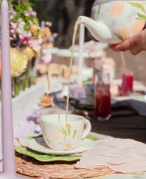Pouring tea from a floral teapot into a matching teacup at a pastel outdoor garden tea-party table with flowers.