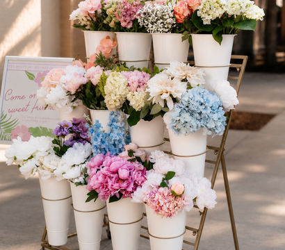 Sunlit outdoor sidewalk flower cart with white cone vases holding pastel bouquets of peonies, hydrangeas, roses and baby's breath