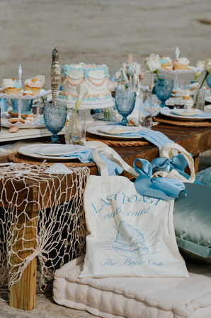 Coastal-chic seaside bridal dessert table with pastel shell-decor cake, cupcakes, blue goblets and nautical netting on a rustic wooden table with a tote bag