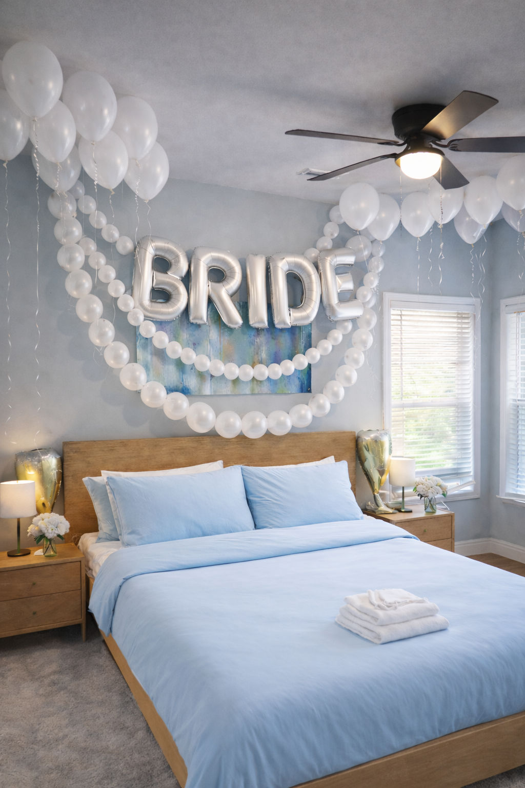 Cozy bridal suite bedroom with light-blue bedding on a wooden bed, silver foil balloons spelling BRIDE and cascading white balloon garlands, ceiling fan, bedside tables with lamps and flowers, and neatly folded towels on the bed.