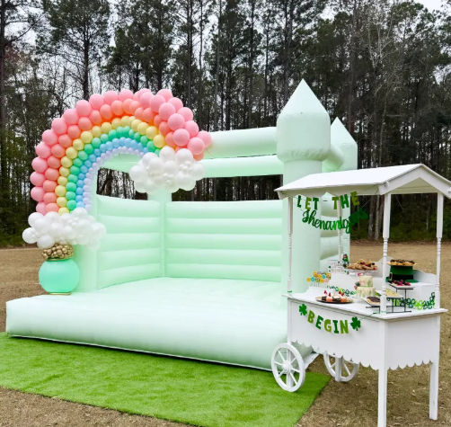 Outdoor pastel mint inflatable castle bouncy house with rainbow balloon arch and white dessert cart on green lawn, pine tree backdrop — playful kids party setup