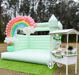 Outdoor pastel mint inflatable castle bouncy house with rainbow balloon arch and white dessert cart on green lawn, pine tree backdrop — playful kids party setup