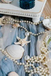 Beach-themed flatlay of pearl necklaces, seashells and a starfish on soft blue tulle beneath a white wooden stand and blue glass jar, coastal table decor.