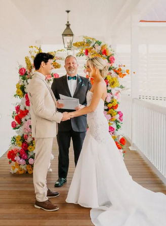 Sunlit wedding on a covered wooden deck: bride in a strapless white gown and groom in a beige suit hold hands under a vibrant floral arch while an officiant conducts the ceremony near a white railing.