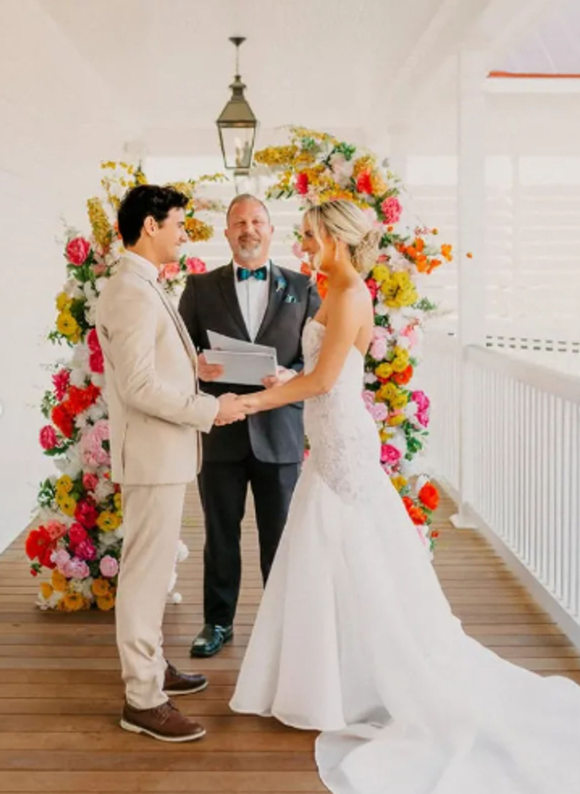 Sunlit wedding on a covered wooden deck: bride in a strapless white gown and groom in a beige suit hold hands under a vibrant floral arch while an officiant conducts the ceremony near a white railing.