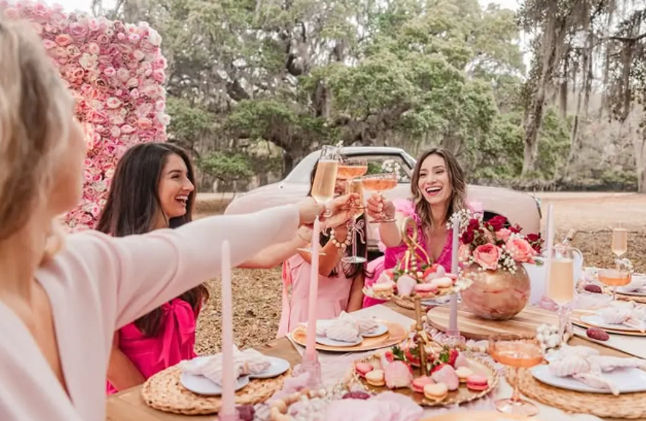 Friends toasting with champagne at a pink-themed outdoor garden picnic, floral wall backdrop, macaron tower, candlelit table and vintage car in an oak grove