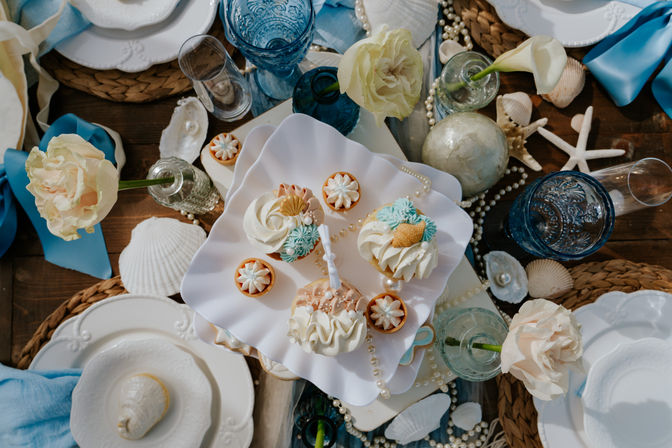 Coastal wedding dessert table with frosted cupcakes and mini tarts on a ruffled white stand, blue vintage glassware, pale roses, pearl strands and seashells on woven placemats — beachy, elegant tablescape