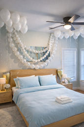Modern bedroom decorated for a celebration: light blue bedding on a natural wood bed, cascading white balloon garlands and floating white balloons from the ceiling, bedside tables with lamps and flowers, ceiling fan and sunlit windows with blinds.