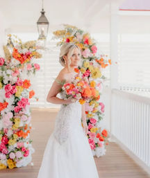 Bridal portrait on a sunlit white-shuttered porch: bride in a lace wedding gown holding a vibrant coral, pink and yellow bouquet beside a colorful flower arch