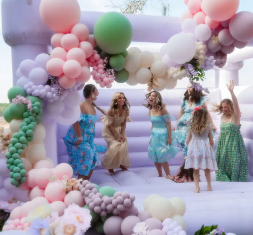 Women and girls laughing and jumping on a pastel lavender bouncy castle decorated with pink, green, and cream balloon garlands and flower accents at an outdoor garden party.