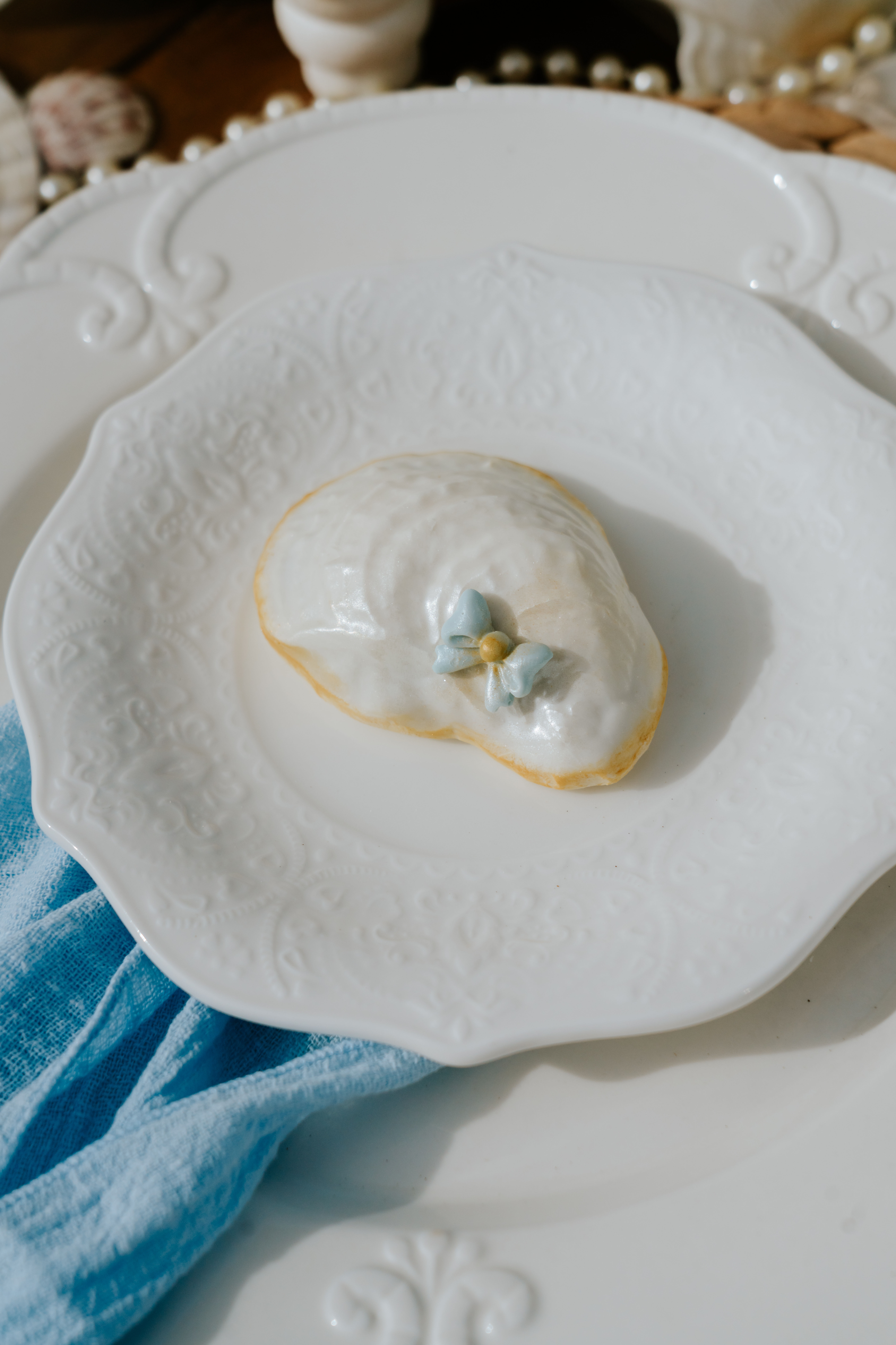 Close-up of a single glazed pastry topped with a tiny blue sugar flower on an ornate white plate, styled with a turquoise linen for an elegant dessert photo.