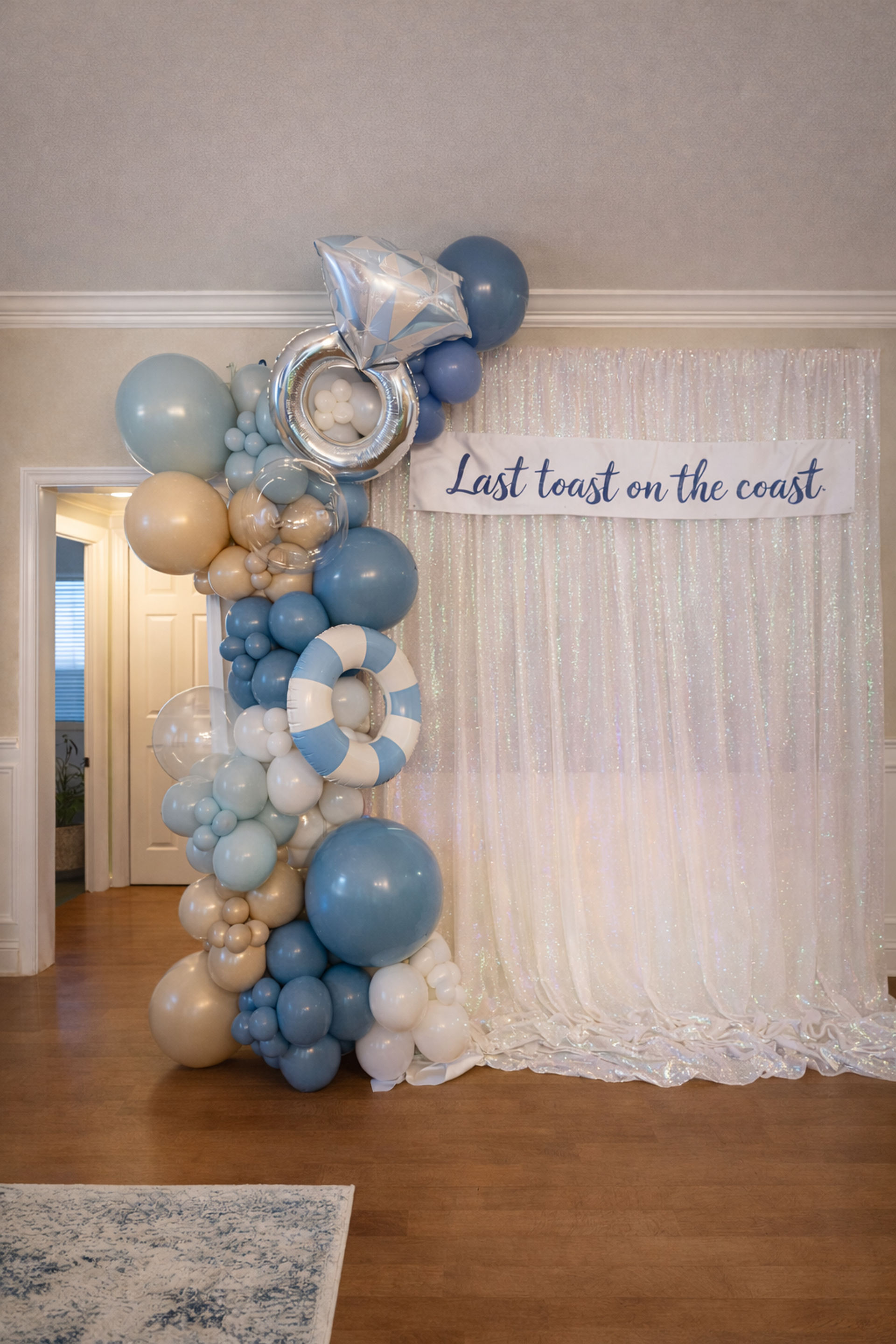 Coastal celebration backdrop with blue, gold and white balloon arch, silver foil ring and striped lifebuoy in front of a shimmering white curtain and banner reading 'Last toast on the coast'.