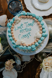 Top-down view of a beach-themed celebration cake reading "Last Toast on the Coast", decorated with seafoam blue and peach frosting swirls, edible seashells and sugar pearls on a seaside table setting