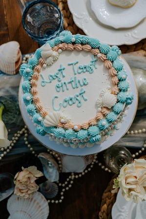 Top-down view of a beach-themed celebration cake reading "Last Toast on the Coast", decorated with seafoam blue and peach frosting swirls, edible seashells and sugar pearls on a seaside table setting