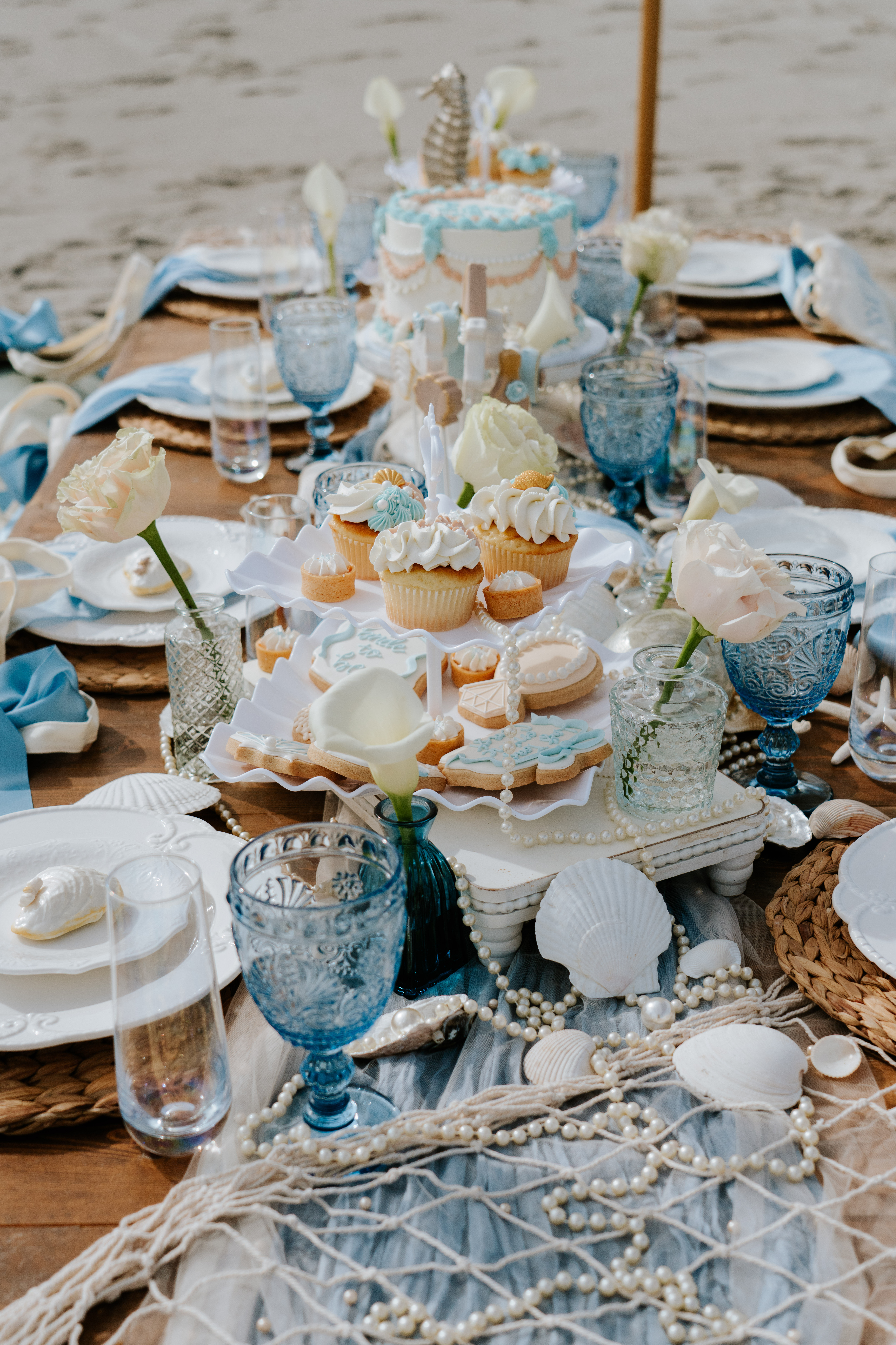 Coastal-chic beachside dessert table with pastel cupcakes and iced cookies on a tiered stand, blue goblets, pale roses, seashells and pearl garlands on woven chargers by the sand