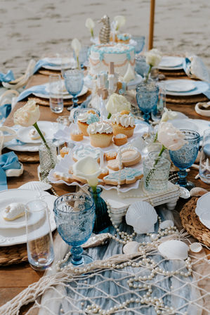 Coastal-chic beachside dessert table with pastel cupcakes and iced cookies on a tiered stand, blue goblets, pale roses, seashells and pearl garlands on woven chargers by the sand